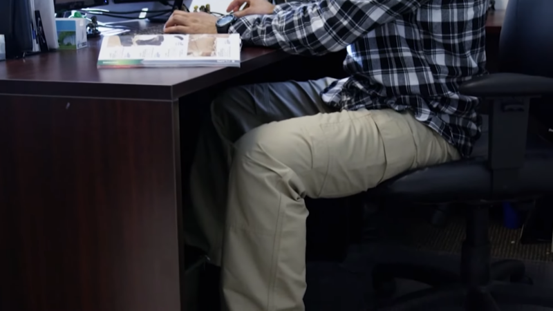 A Person Sitting at An Office Desk Wearing Tactical Pants and A Plaid Shirt