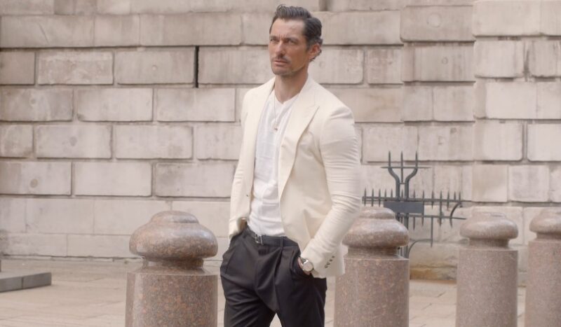 A man in a cream-colored suit stands before a light beige brick wall